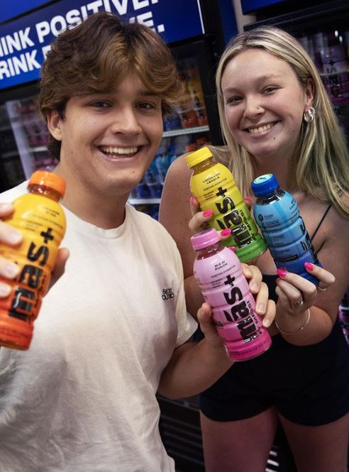 Two smiling people hold all four flavors of the Más+ by Messi range, featuring bottles in yellow, pink, orange, and blue. In the background, a refrigerated display showcases additional bottles alongside a "THINK POSITIVE. DRINK POSITIVE" sign.