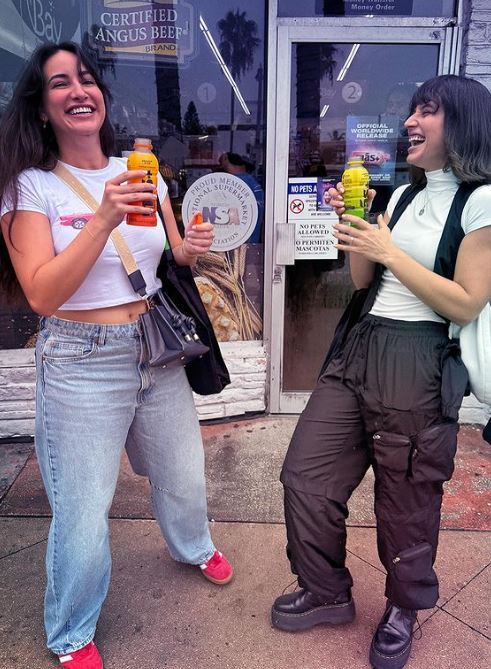 Two girls stand in front of a store holding Más+ by Messi drinks while laughing.