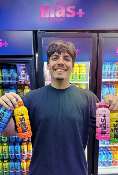 A person holds three bottles of Más+ by Messi in front of a refrigerator.