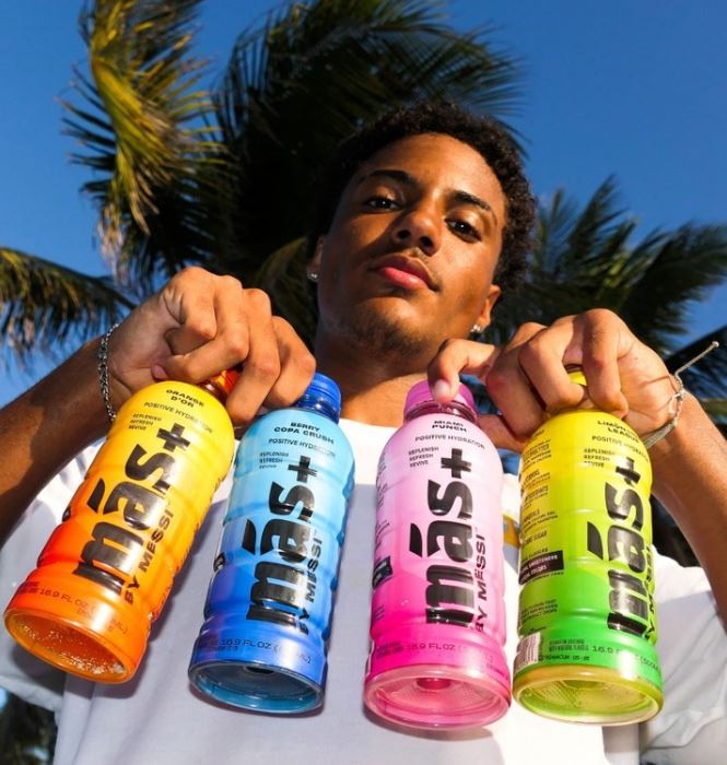 A young man holds four colorful bottles of Más+ by Messi drinks in yellow, pink, blue, and orange.