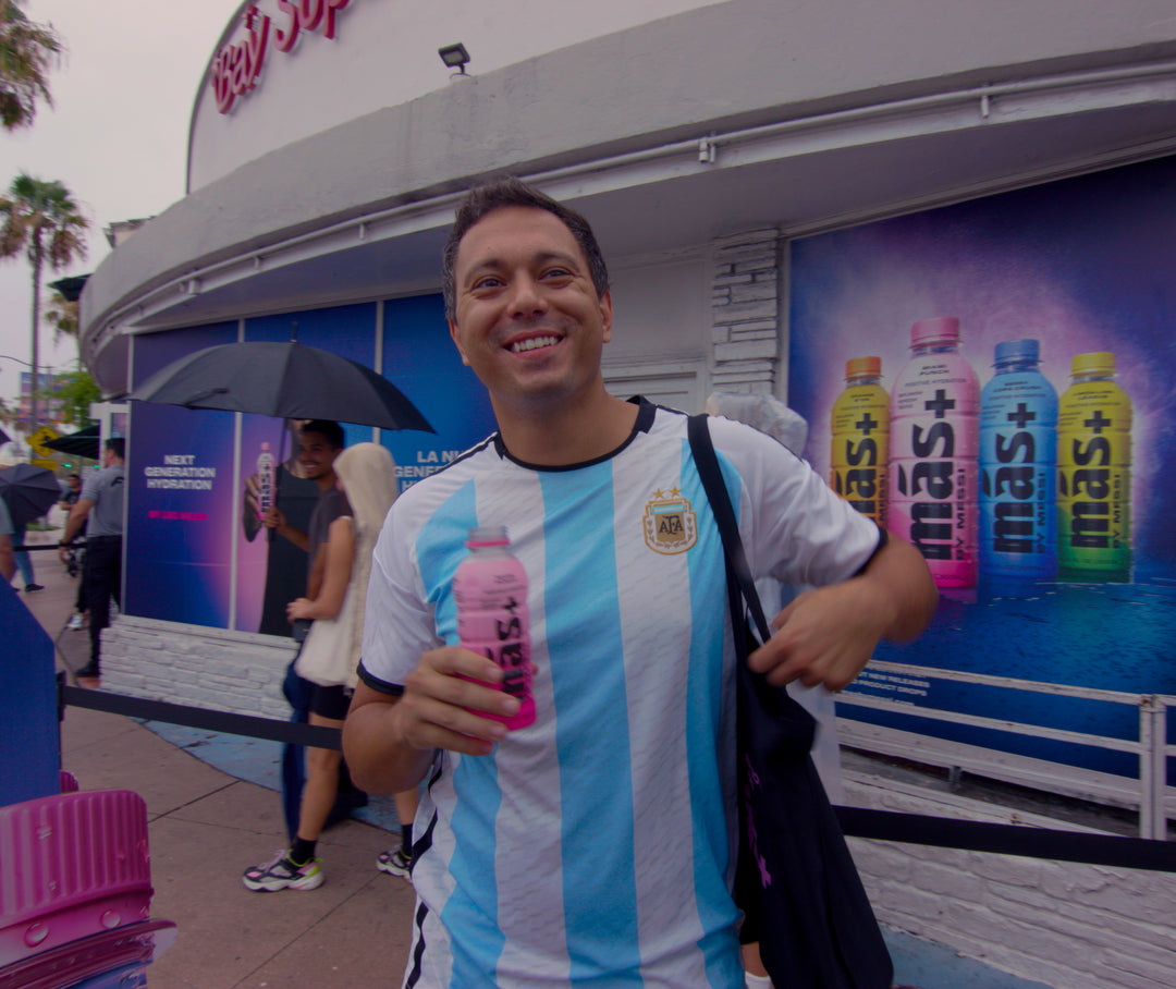 A man wearing an Argentina football T-shirt holding a Más+ by Messi bottle in Miami Punch flavor.