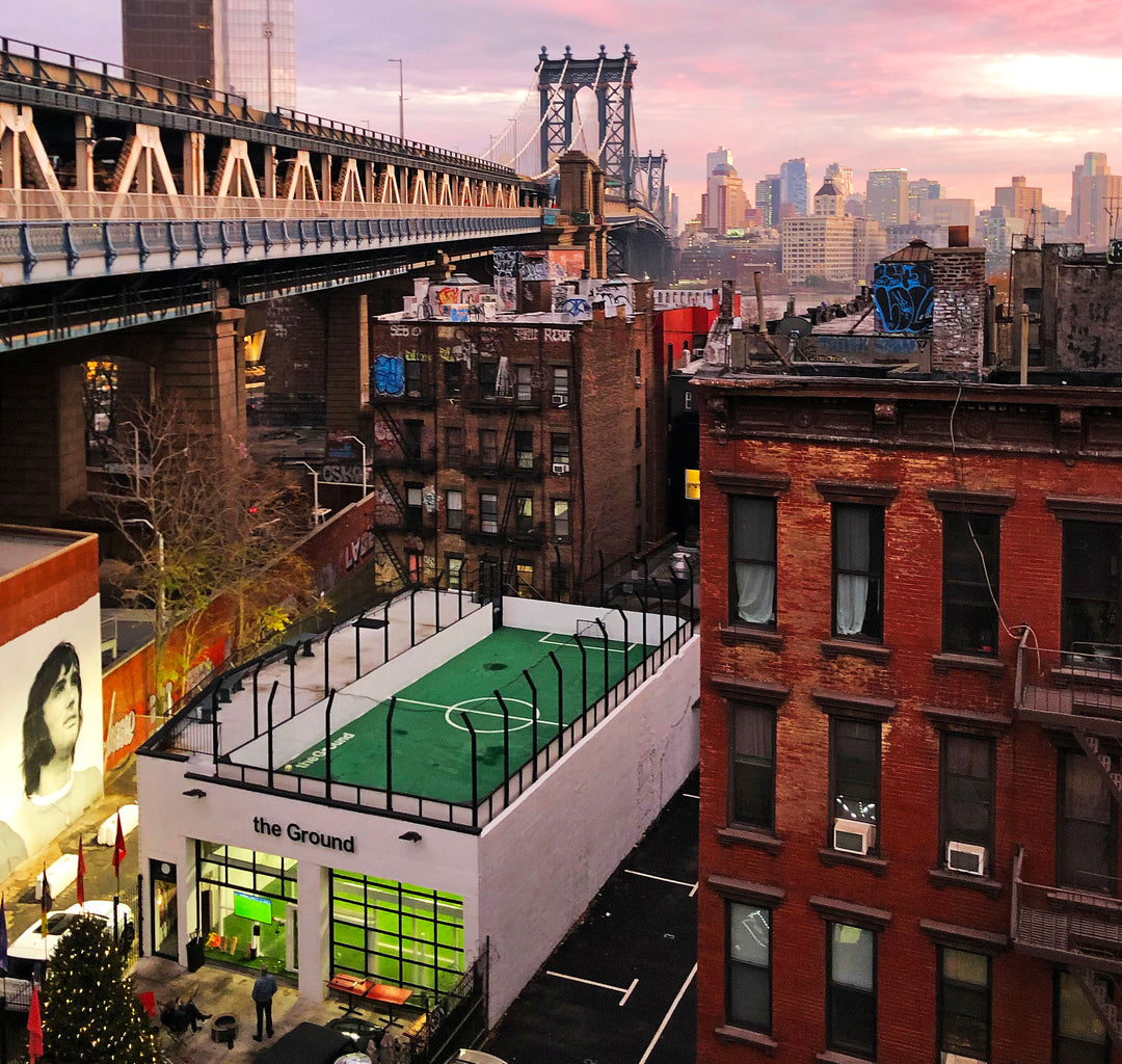 The "The Ground" building in a lively urban area features a fenced rooftop soccer field, surrounded by city views and a large mural on a nearby wall. A man watches TV outdoors.