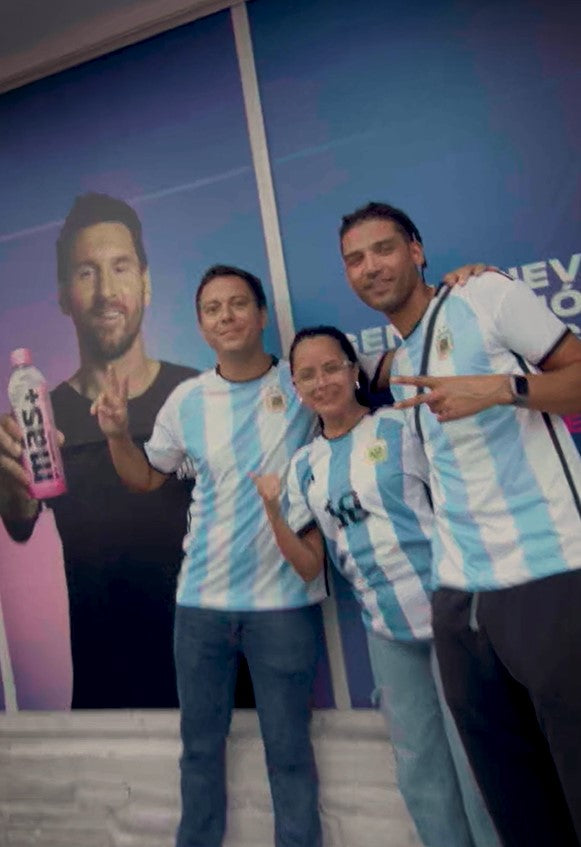 Three people wearing Argentina t-shirts stand in front of a poster of Leo Messi