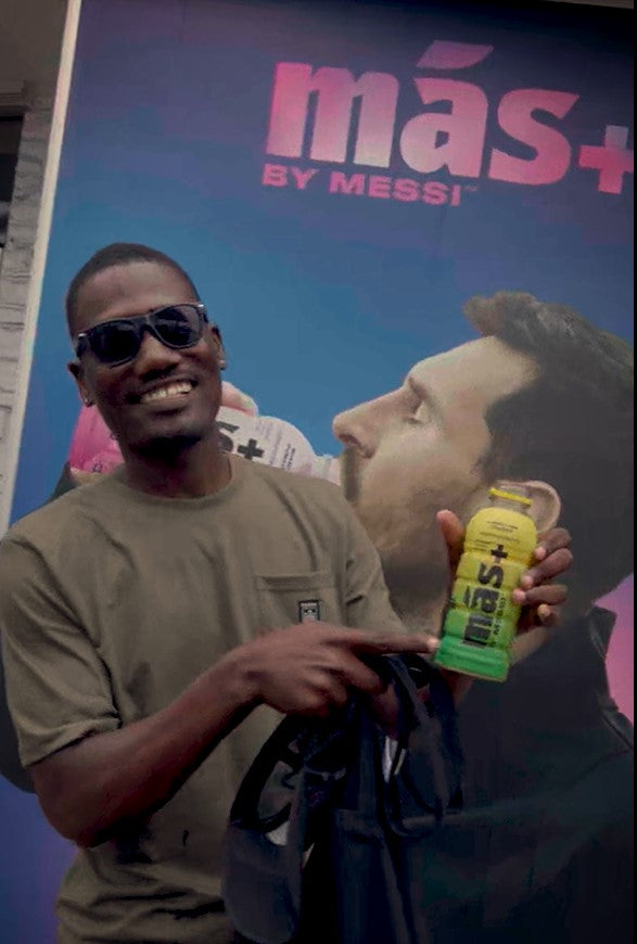 A person smiles and points at a yellow bottle of Más+ by Messi in their hand. Behind them is an advertisement featuring Leo Messi with the text "Más+ by Messi".