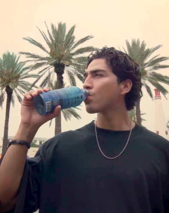 A person drinks a Más+ by Messi Copa Crush hydrating drink in an outdoor setting, featuring tall palm trees and a lighthouse in the background. They are wearing a dark shirt and a necklace.